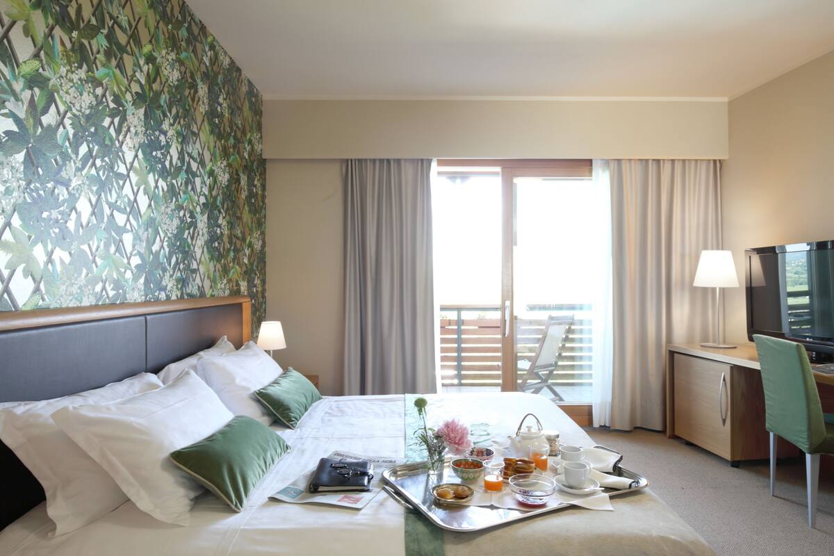 A hotel room with a bed, a desk with a chair, a TV, and a balcony with curtains.