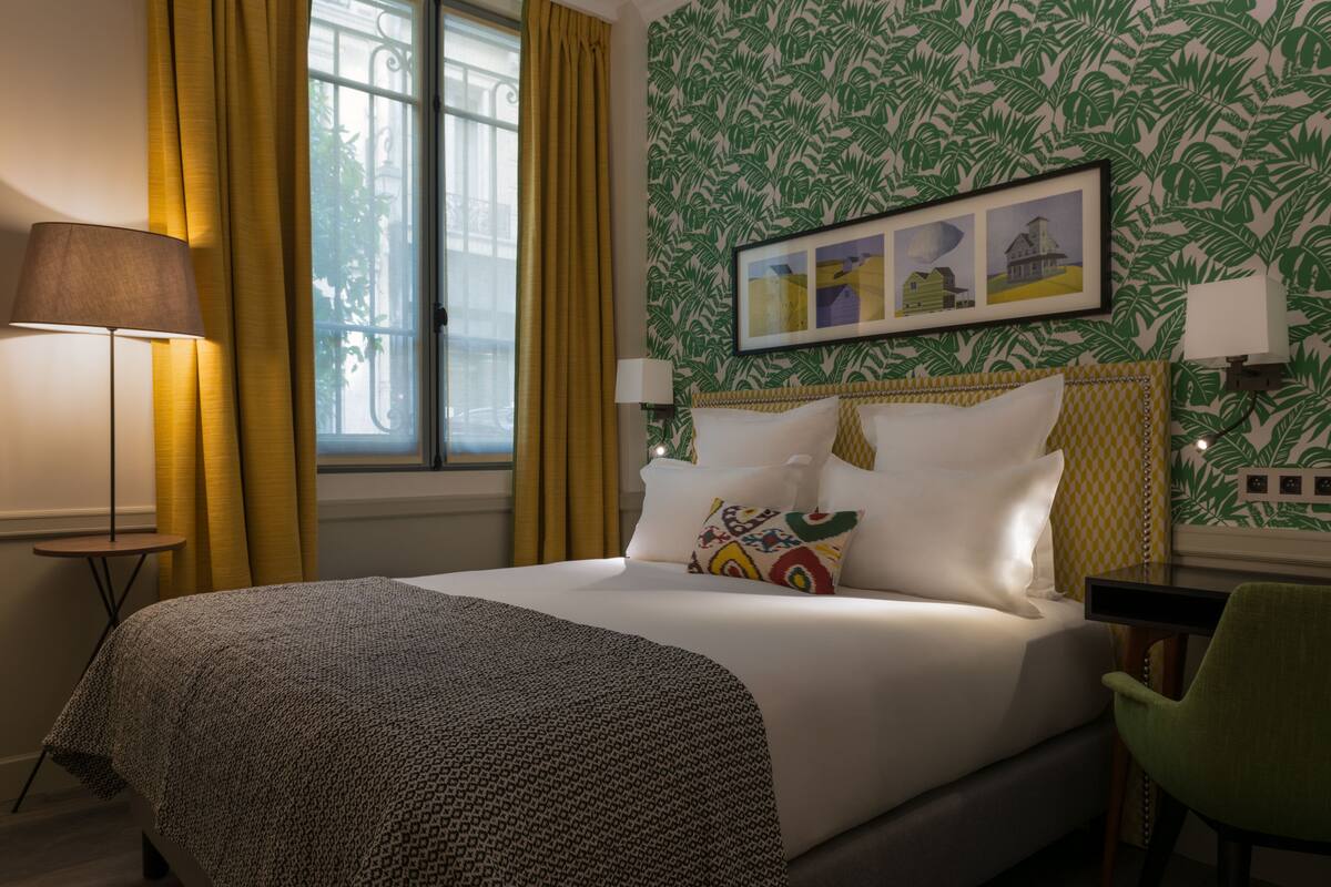 A bed with patterned bedding, a bedside lamp, a framed picture, and a window with curtains.