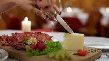 A hand using a knife to cut a block of cheese on a wooden board, surrounded by various meats and vegetables.