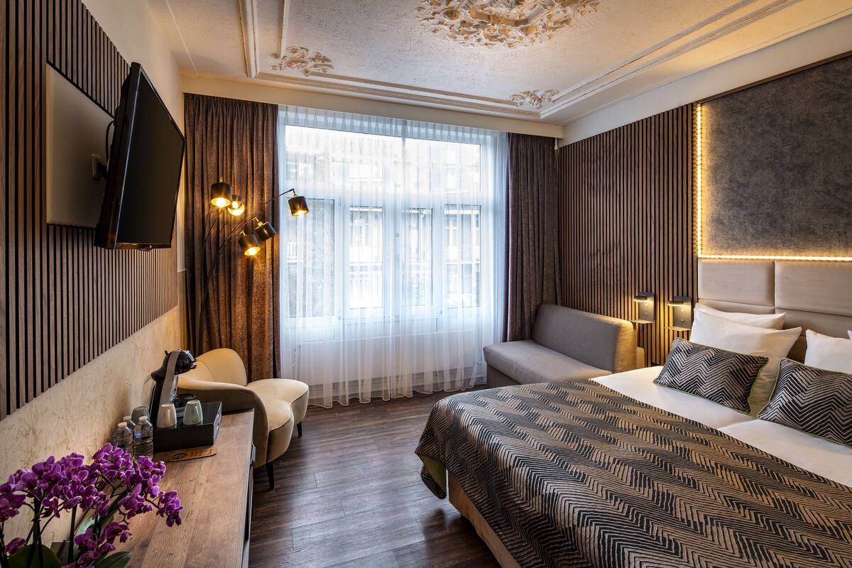 A hotel room with a bed, a sofa, a TV, a desk with a lamp, and a window with curtains.