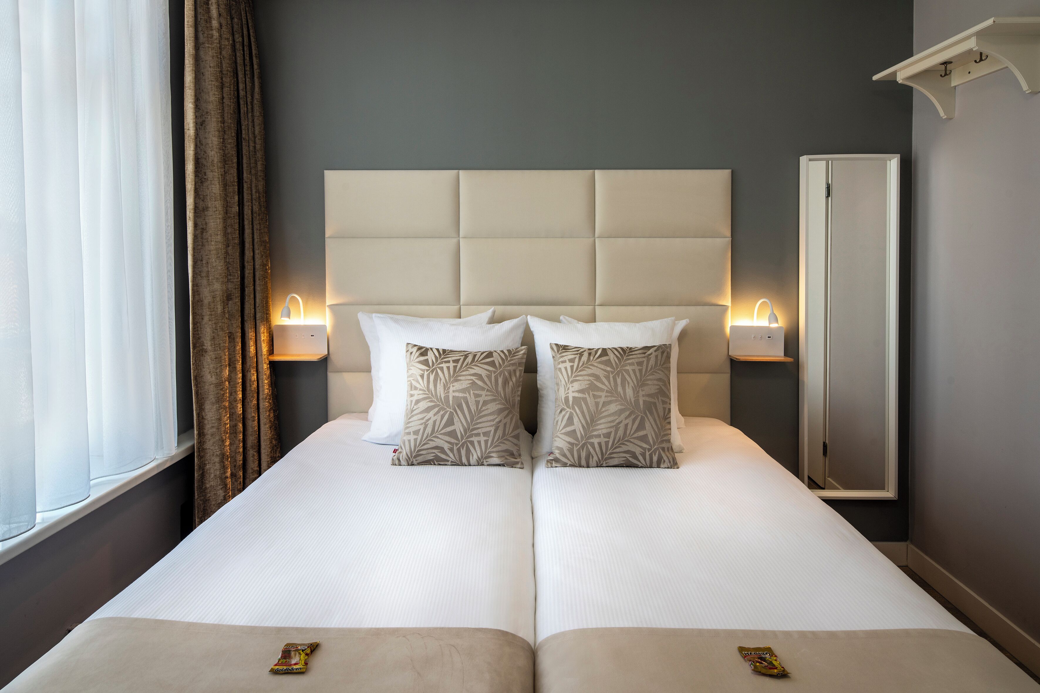 Een tweepersoonsbed met een gestoffeerd hoofdbord, twee bedlampjes en een spiegel aan de muur.