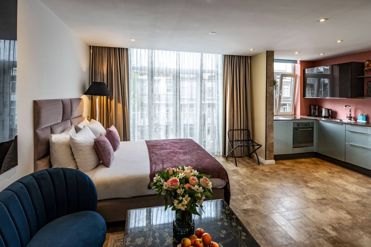 A modern hotel room with a large bed, a sitting area with a sofa and armchair, a dining table with fruit, and a view of the city through large windows.