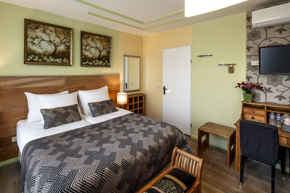 A bedroom with a large bed, a desk, a chair, a small table, and a TV on the wall.