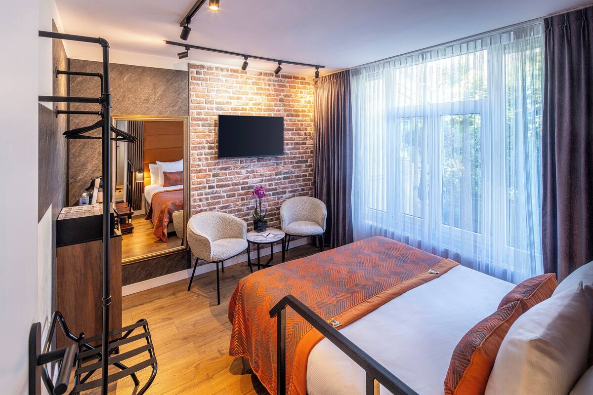 A hotel room with a brick wall, a bed with orange bedding, a TV, a desk with a chair, and a window with curtains.