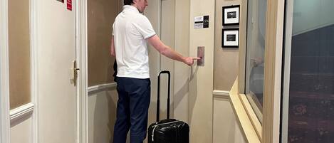 A man in a white shirt and dark pants standing in a hallway with a black suitcase, approaching an elevator with a digital display and framed pictures on the wall.