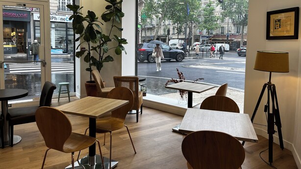 A modern cafe interior with wooden tables and chairs, large windows offering a view of the street, and indoor plants.