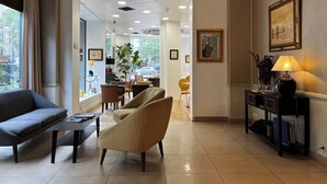 A hotel lobby with a sofa, armchair, and a table with a lamp. There are framed pictures on the wall and a plant on the side.