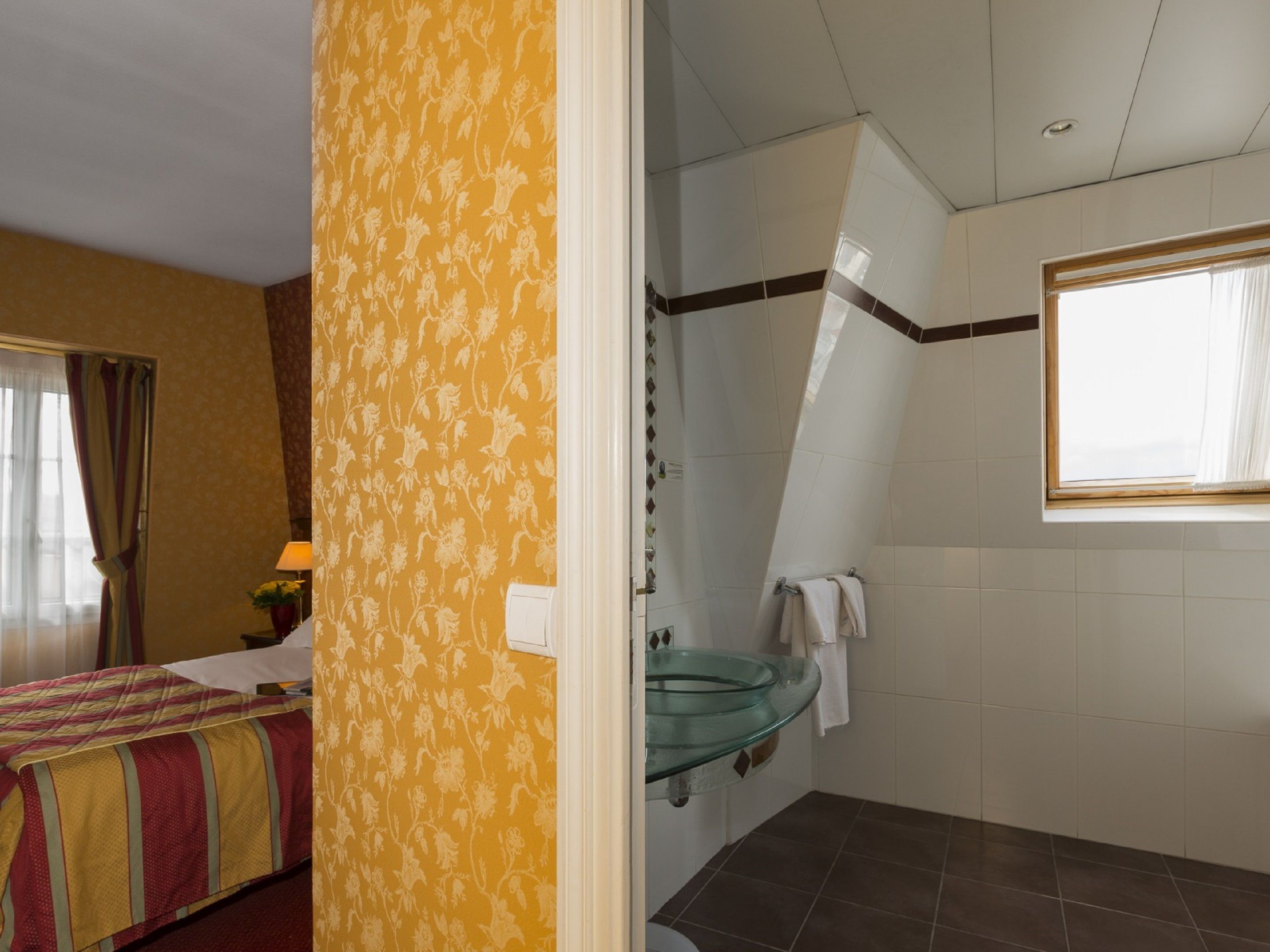 A hotel room with a bed, a bathroom with a glass sink, and a window.