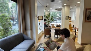 A hotel lobby with a grey sofa, a coffee table with magazines, and a person reading a book. There are framed pictures on the walls and a view of the street outside.