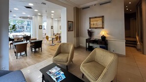 A hotel lobby with a modern design, featuring a reception desk, seating area, and a view of the street through large windows.