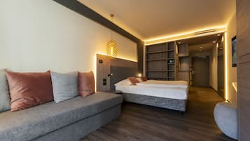 A modern hotel room with a grey sofa, a bed, a small table, and built-in shelving.