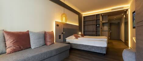 A modern hotel room with a grey sofa, a bed, a small table, and built-in shelving.