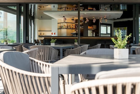An outdoor seating area with wicker chairs and tables, a bar with shelves of bottles, and a large window providing a view of the outdoors.. Ideal Park