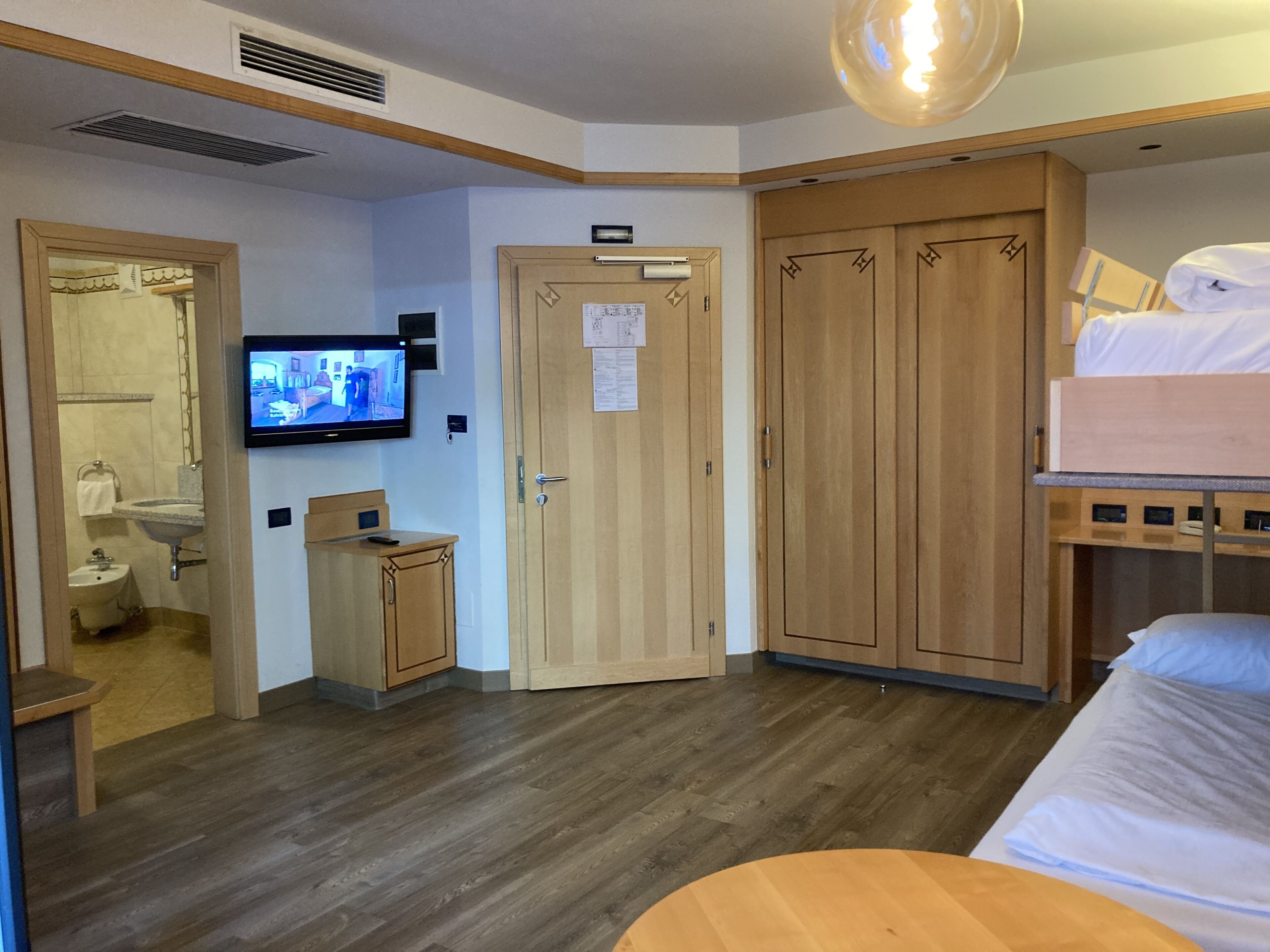 Ein Hotelzimmer mit Holzboden, einem an der Wand montierten Fernseher, einem Holzschrank, einem kleinen Holzschränkchen und einem Bett mit weißer Matratze und Kopfkissen.