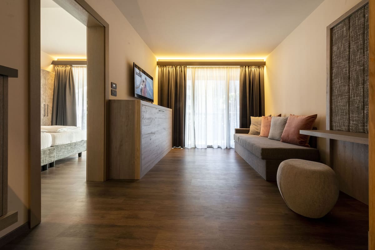 Una habitación de hotel moderna con piso de madera, un sofá con cojines, un televisor sobre un mueble de madera y un amplio ventanal con cortinas.