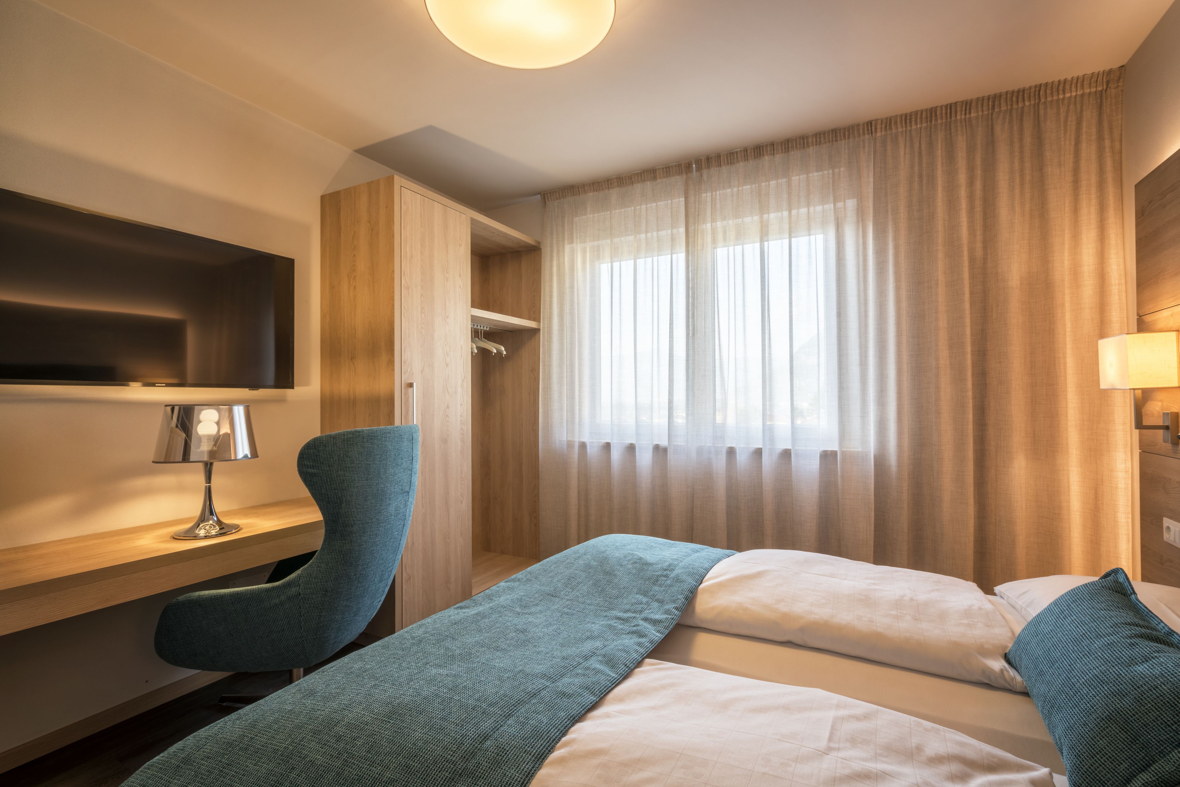 A hotel room with a bed, a desk, a chair, a TV, and a window with curtains.
