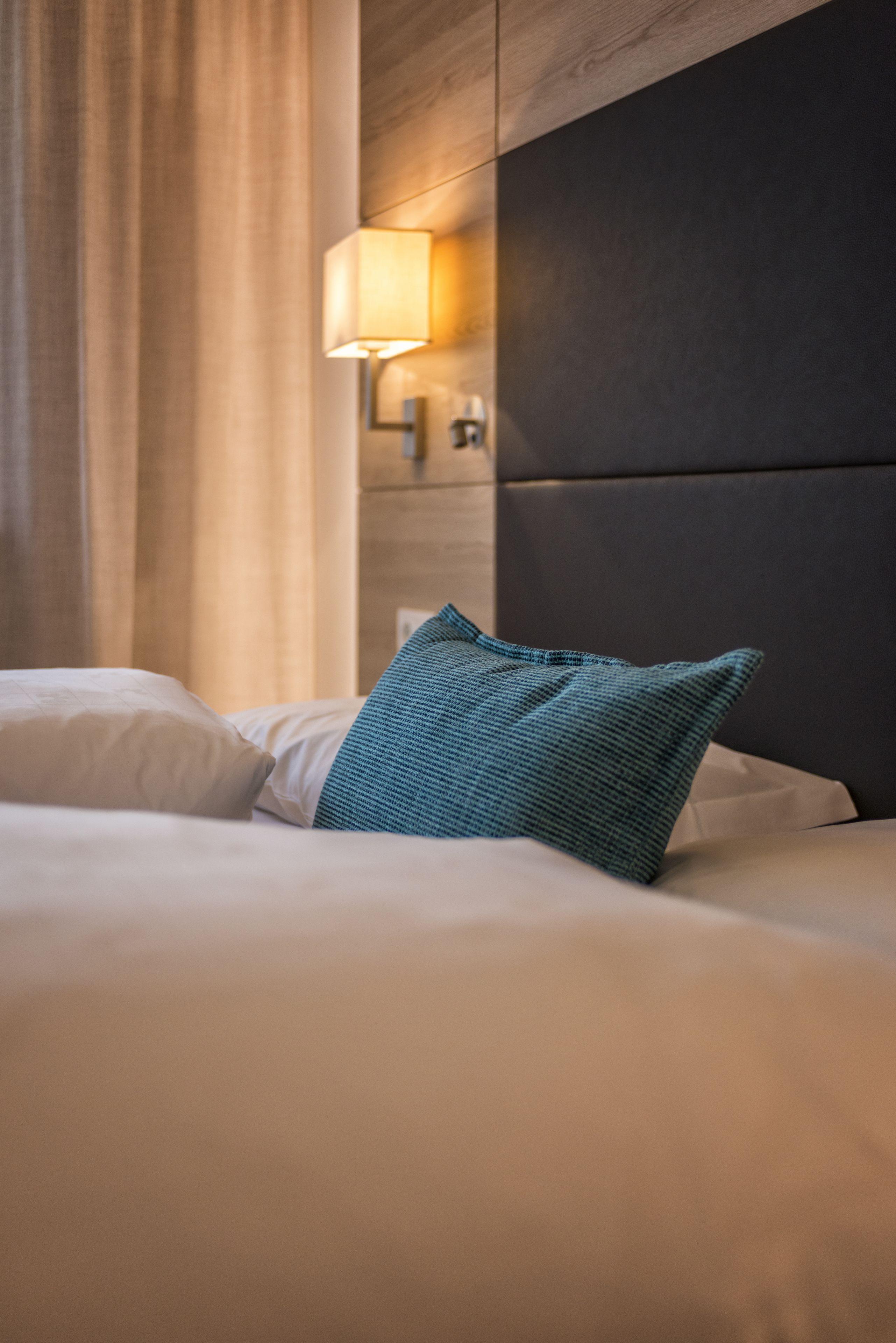 Una habitación de hotel con una cama, un almohadón verde azulado, una lámpara fijada a la pared y una ventana con cortinas.