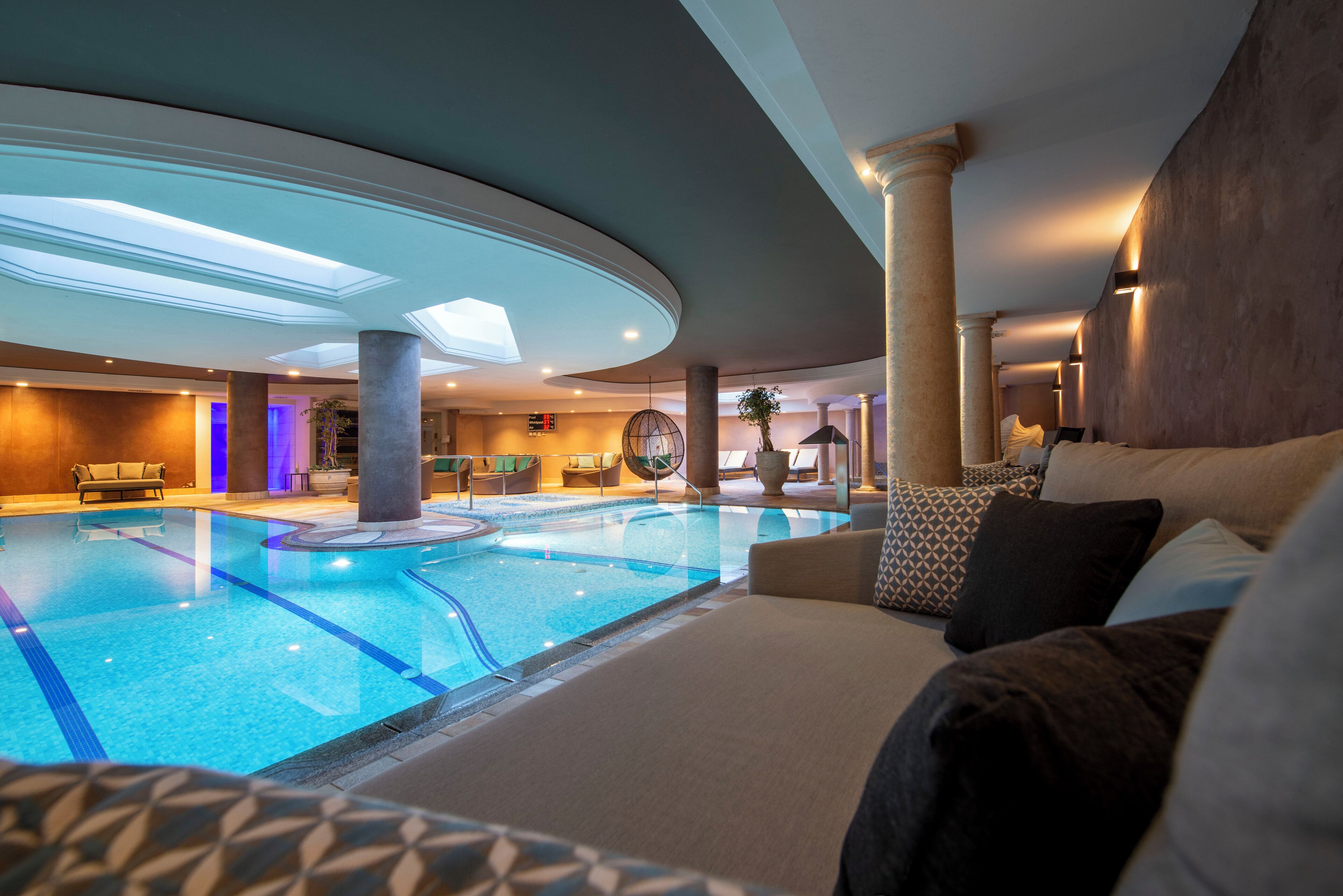 Piscina interna com áreas de descanso, teto com claraboias e design de interiores moderno.