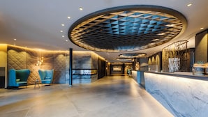 Un moderno vestíbulo de hotel con una recepción de mármol, un área de descanso con sillones azules y un diseño de techo geométrico.