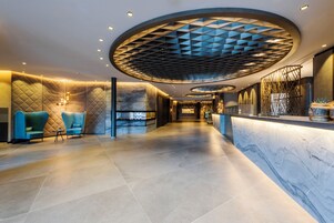 Een moderne hotellobby met een marmeren receptiebalie, een zithoek met blauwe fauteuils en een geometrisch plafondontwerp.
