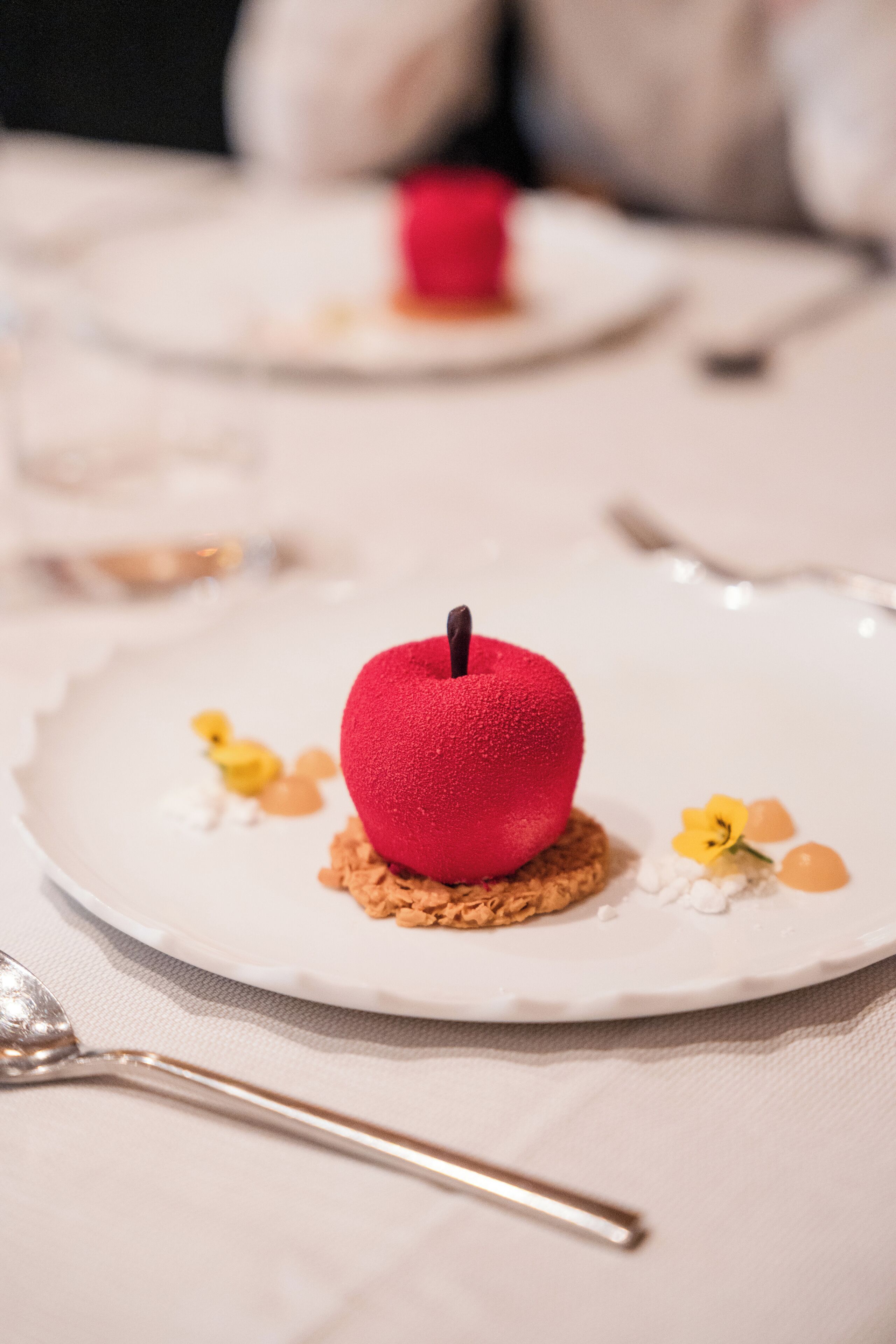 Un plato de postre con una manzana roja como elemento central, una cuchara y un plato blanco al fondo.
