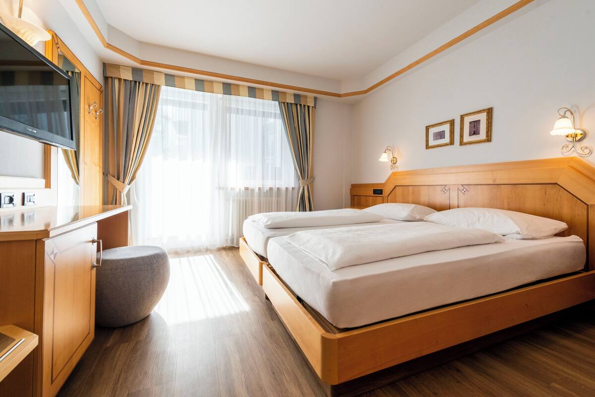 Habitación de hotel con cama de madera, un otoman redondo, televisor y ventana con cortinas.