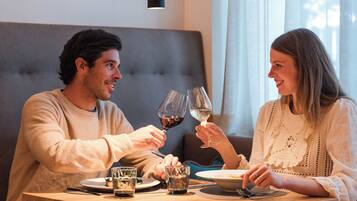 Una pareja sentada en una mesa de madera, cada uno con una copa de vino, en una habitación con un sofá tapizado en gris y una lámpara colgante.