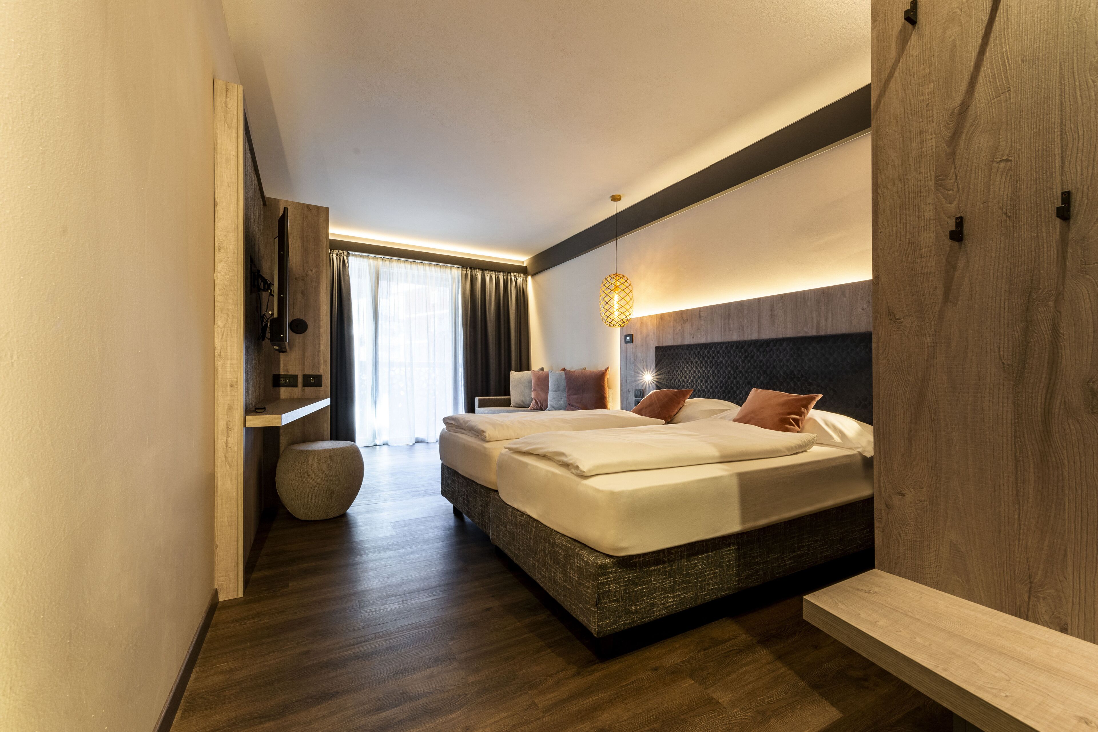 Quarto de hotel moderno com cama grande, mesa de cabeceira, TV integrada e uma ampla janela com cortinas.