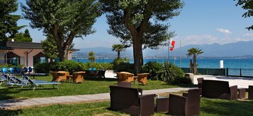 Un área de estar al aire libre frente al mar, con sillones y mesas, con vistas a la costa y montañas a lo lejos.
