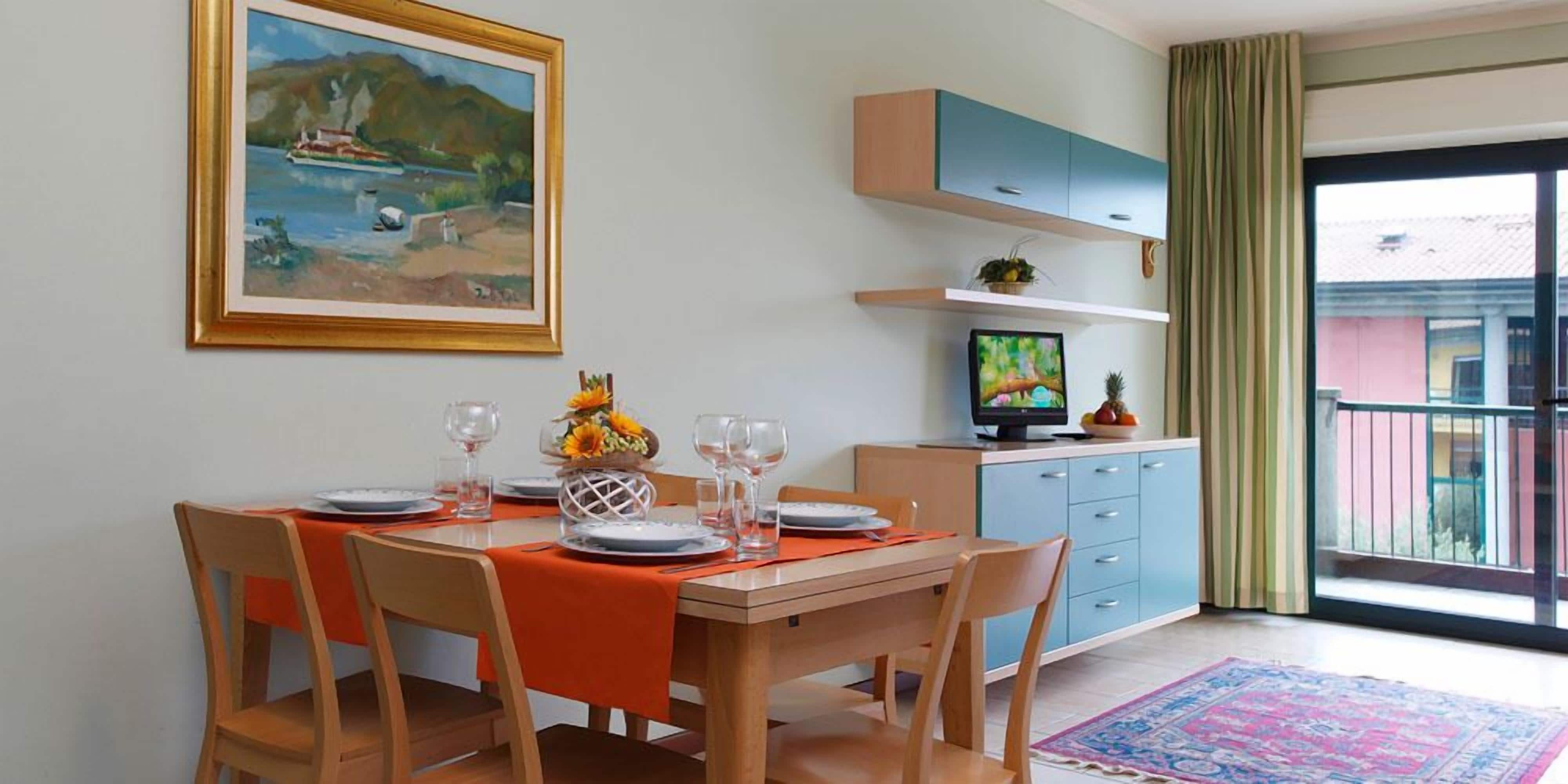 A dining area with a table set for four, a painting on the wall, a television, and a view of the outside balcony.