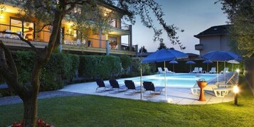Un área de piscina con sillas de descanso y sombrillas, rodeada de follaje y un edificio con balcón.
