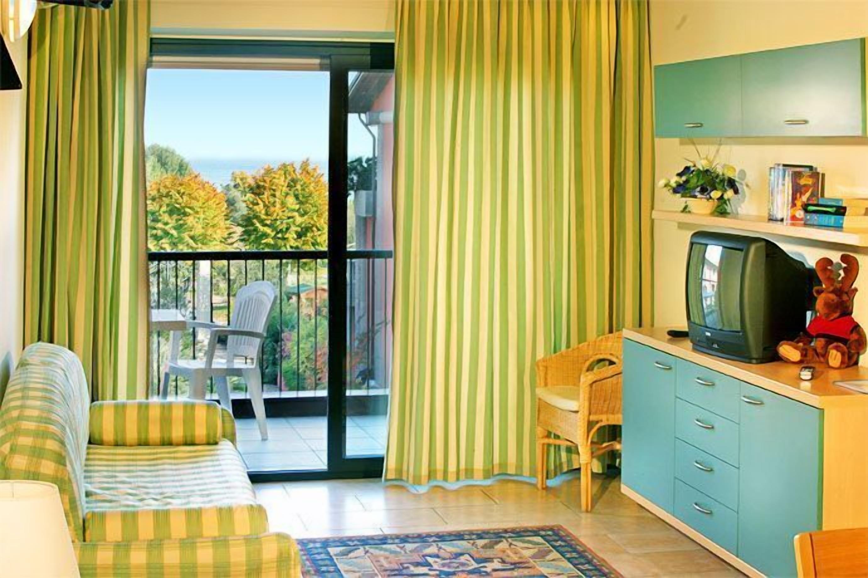 A room with a sofa, a television, a small table, and a cabinet. There is a view of trees and a building through the window.