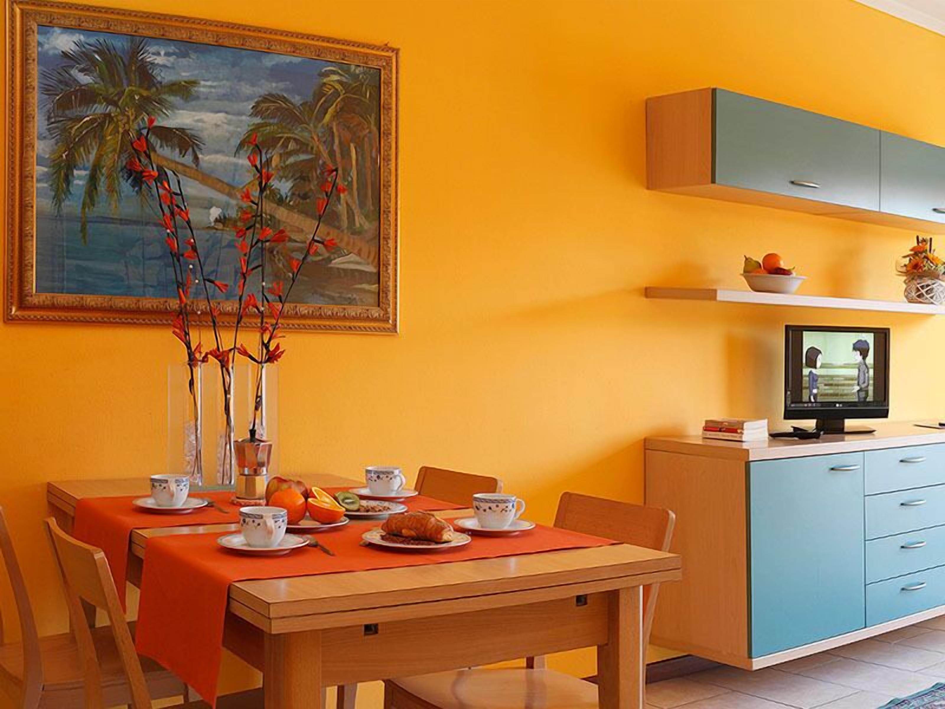 Een eetgedeelte met een ontbijttafel, een schilderij van een tropisch landschap, en een televisie op een standaard.