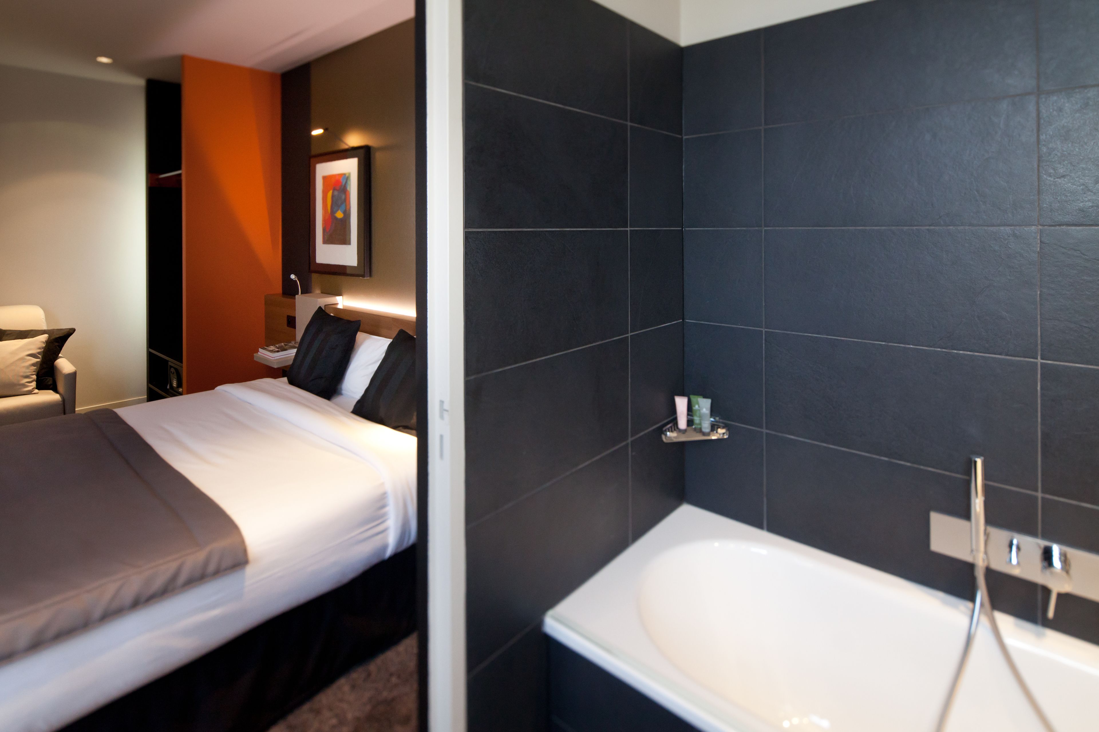 A modern hotel room with a neatly made bed, a large bathtub, and a dark tiled wall.