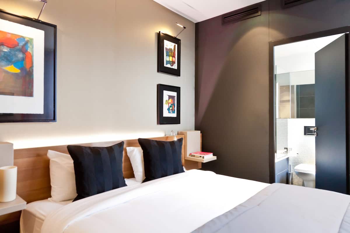 Habitación de hotel con una cama grande, dos cuadros enmarcados en la pared, una mesita de noche con lámpara y vistas al baño por una puerta abierta.