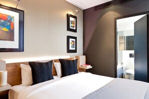 Une chambre d’hôtel avec un grand lit, deux œuvres d’art encadrées au mur, une table de chevet avec une lampe, et une vue sur la salle de bain par une porte ouverte.