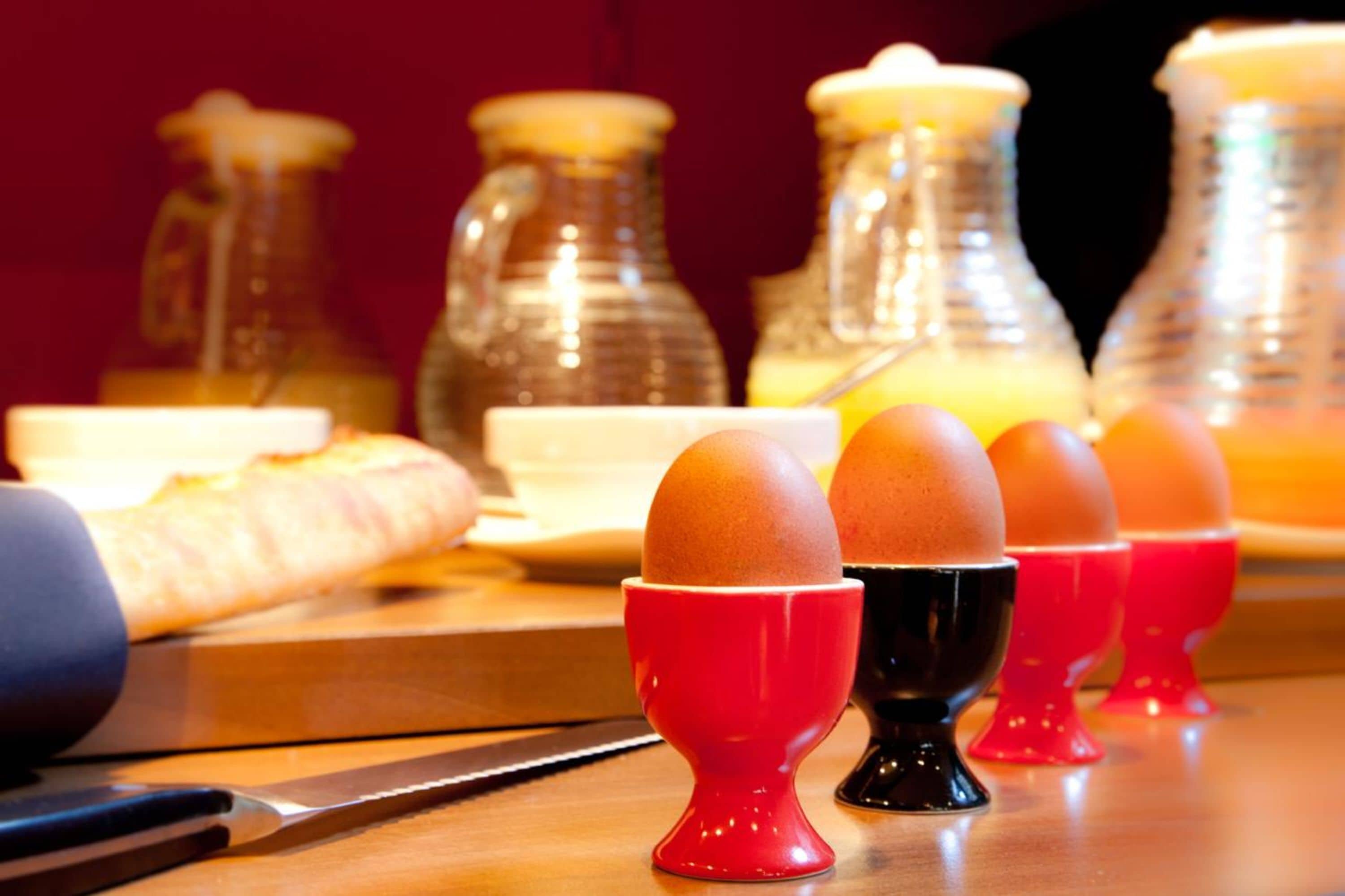 Disposição para o pequeno-almoço com ovos em chávenas vermelhas e pretas, uma faca e uma gaveta de pão sobre uma mesa de madeira.