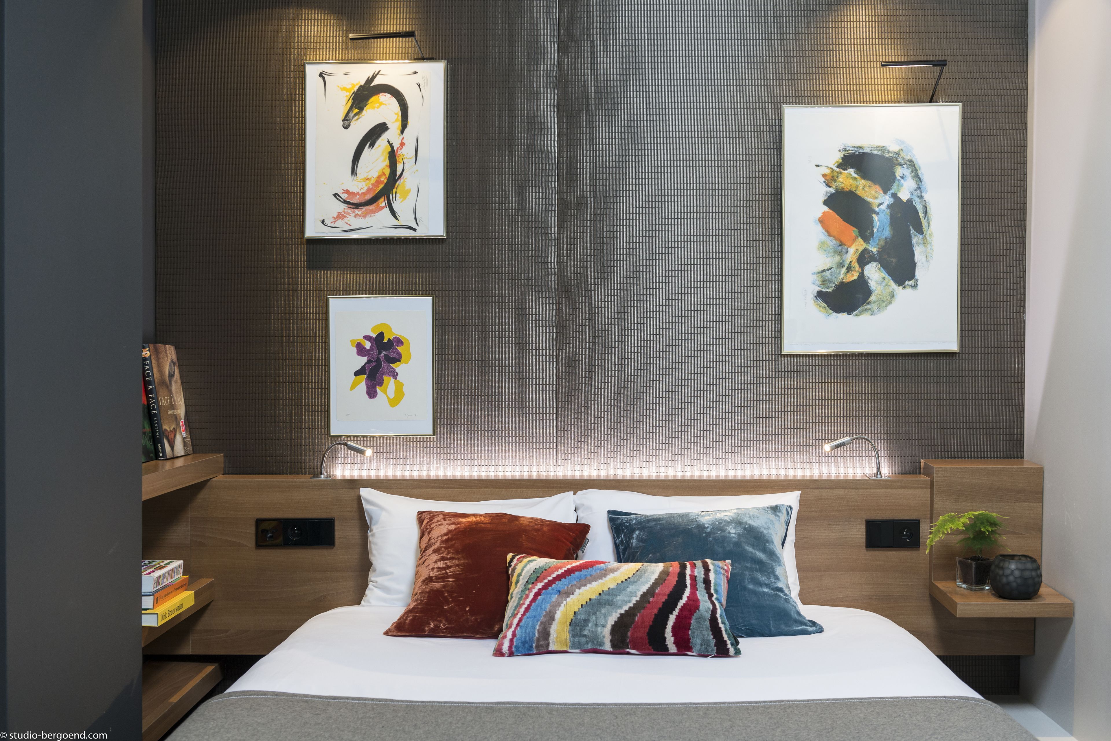 A modern hotel room with a large bed, wooden headboard, and bedside tables with lamps. There are three framed artworks on the wall above the bed.