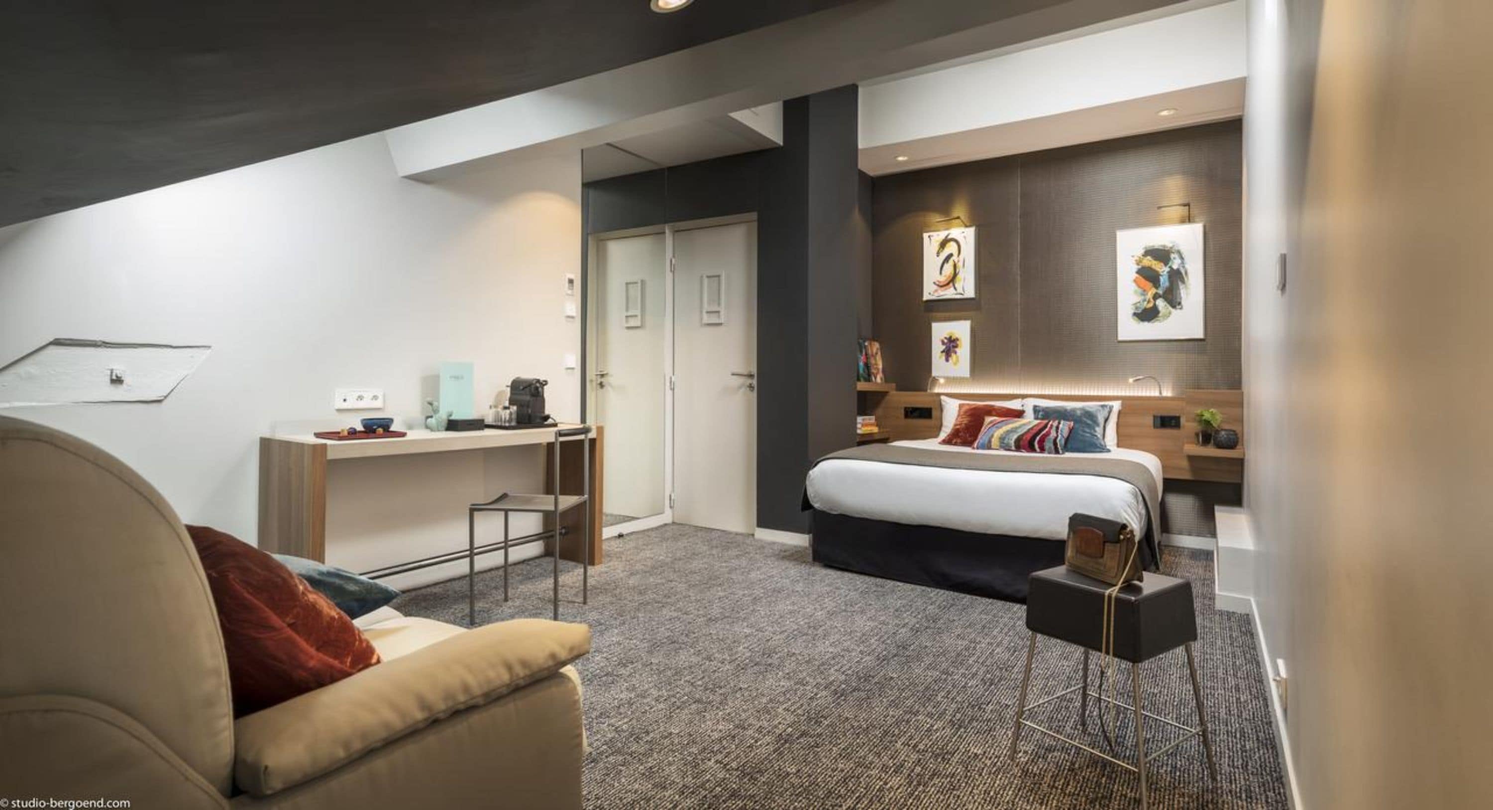 A modern bedroom with a bed, a desk, and a chair. There is a wardrobe and artwork on the wall.