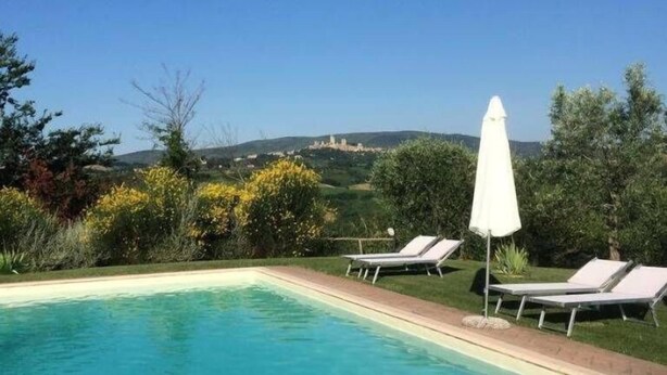 Piscina azul cristalina con sillas de descanso y una sombrilla, rodeada de vegetación y una vista distante de una colina con una construcción.