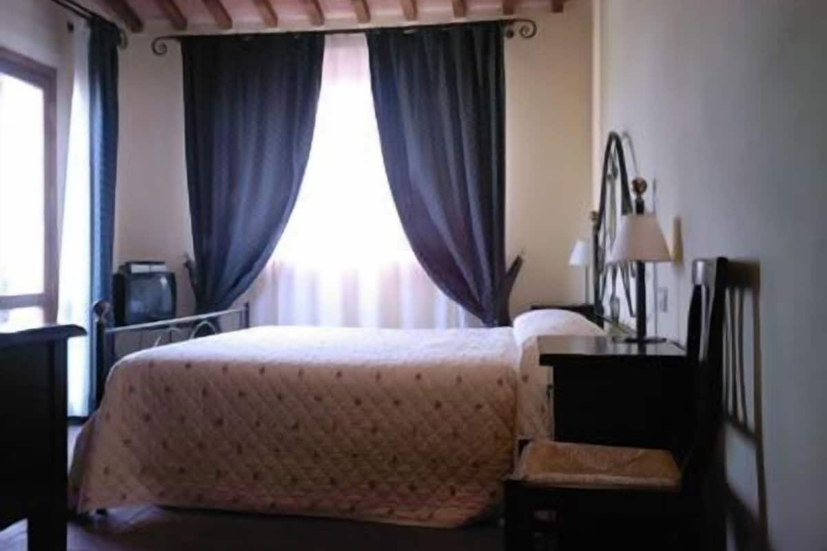 Habitació d'hotel amb llit, escriptori, cadira, làmpada i finestra amb cortines.