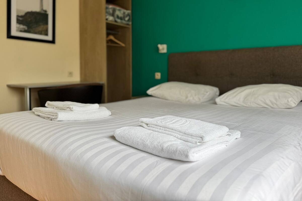A neatly made bed with folded white towels, a wooden wardrobe, and a green wall with a framed picture.