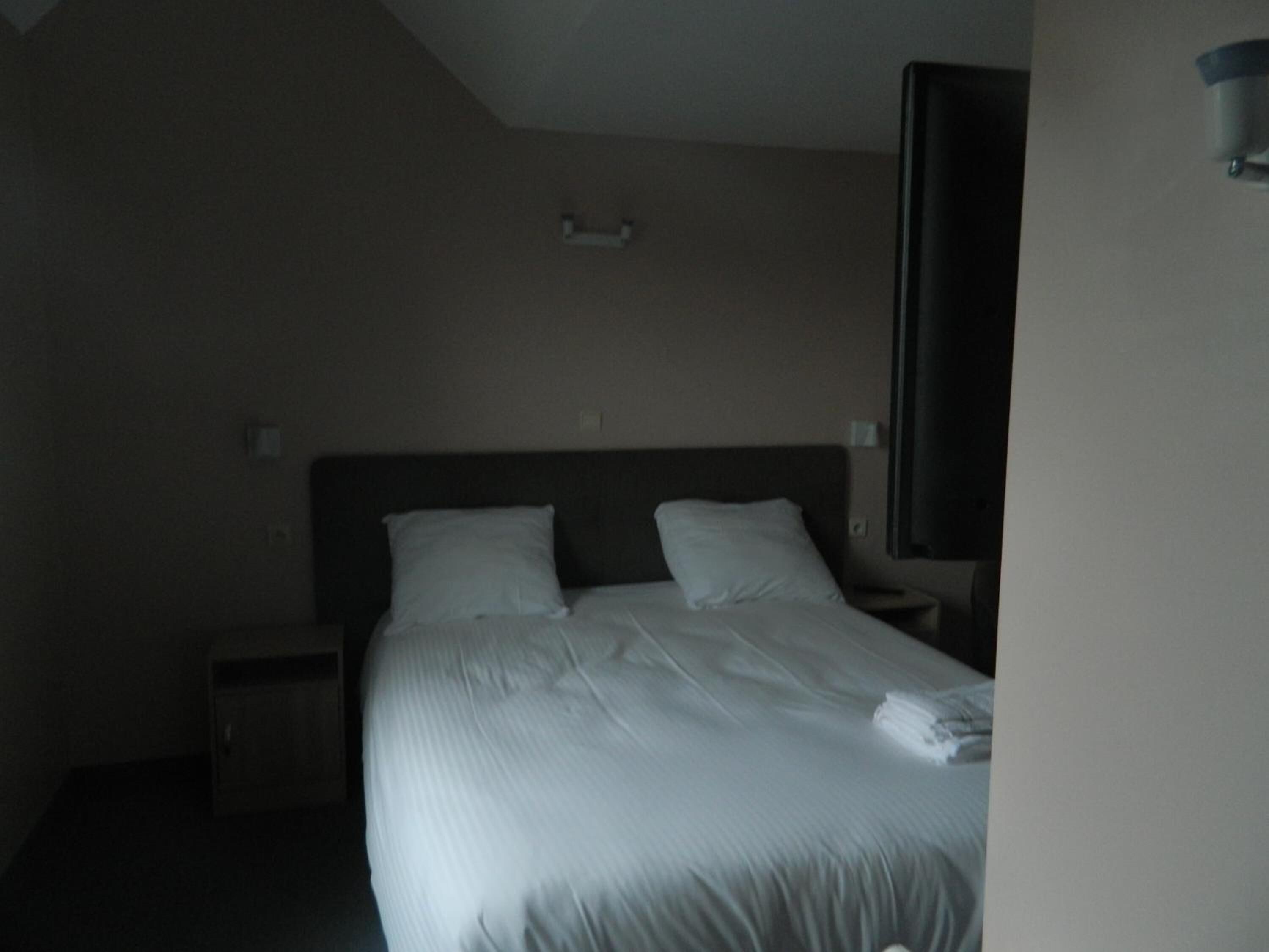 Quarto de hotel com uma cama grande, duas almofadas, uma mesa de cabeceira e televisão fixada na parede.