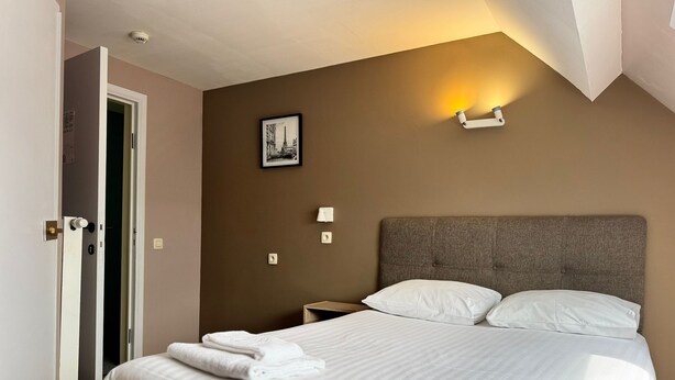 A hotel room with a bed, a nightstand, a wall-mounted lamp, and a door.