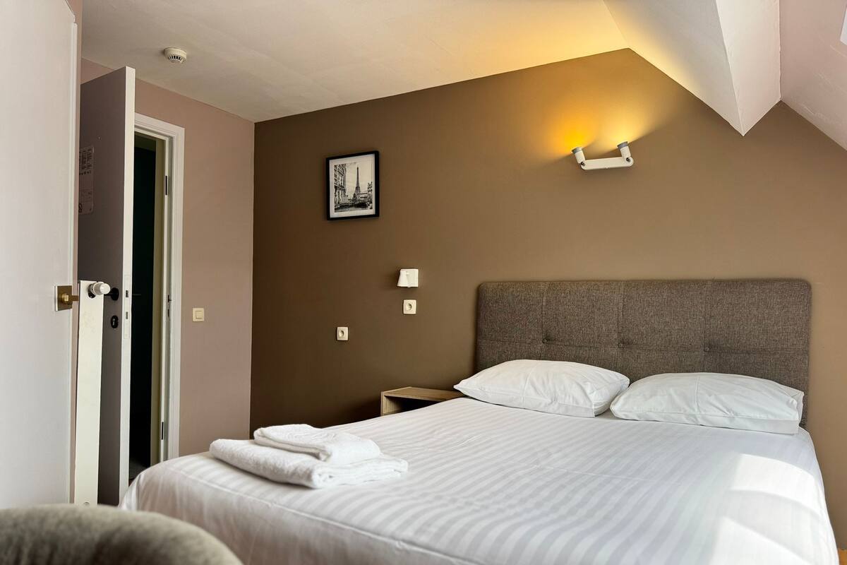 A hotel room with a bed, a nightstand, a wall-mounted lamp, and a door.