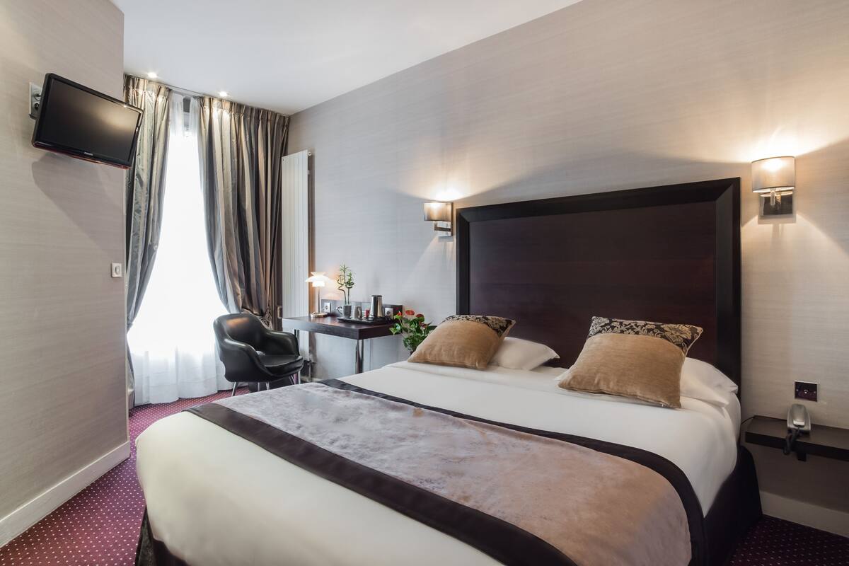 A hotel room with a large bed, a desk with a chair, a flat-screen TV mounted on the wall, and two wall-mounted lights.