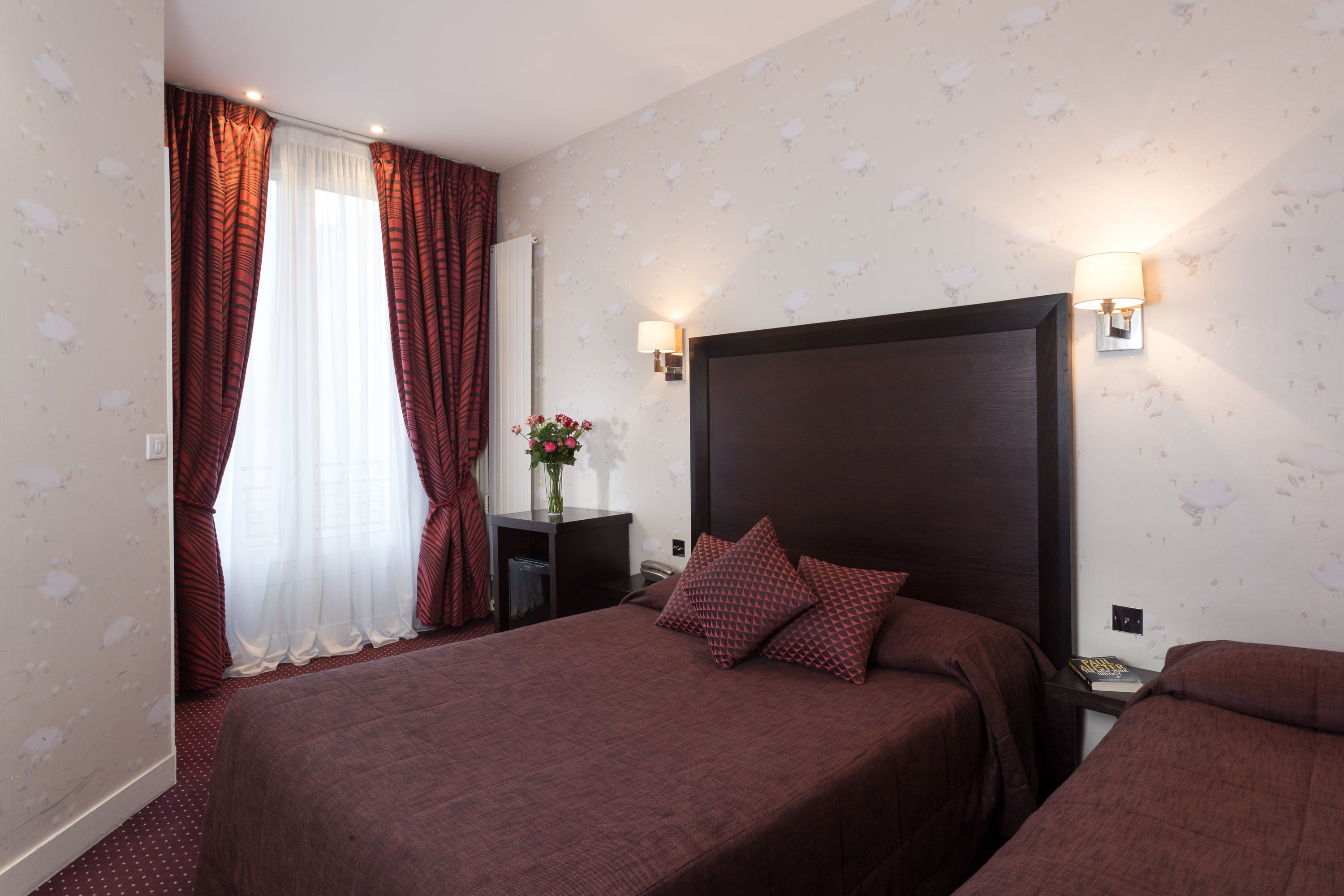 A hotel room with two beds, a nightstand, a lamp, and a window with curtains.