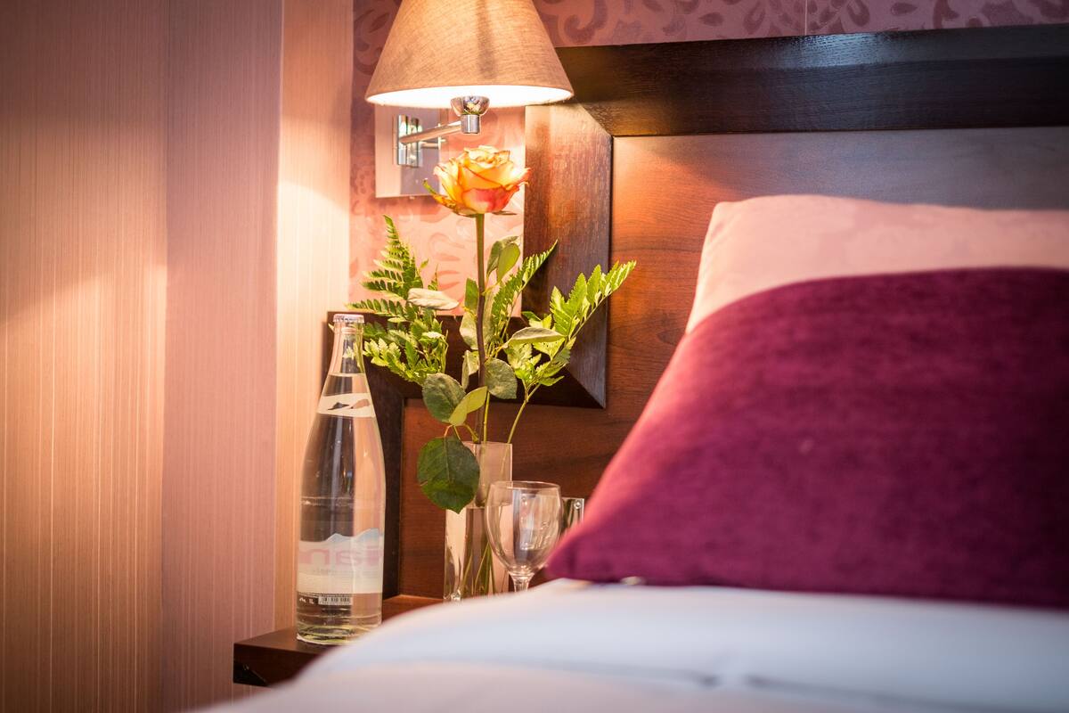 A neatly made bed with a purple pillow, a bedside table with a bottle, glasses, and a vase with a rose.