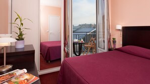 A hotel room with a bed, a desk with a lamp, a chair, a small table with a plant, and a balcony with a view of buildings. - Hotel Virgina (Paris)