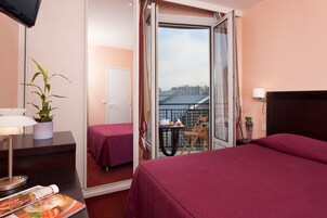 Habitación de hotel con cama, escritorio con lámpara, una silla, una mesita con planta y balcón con vistas a edificios.